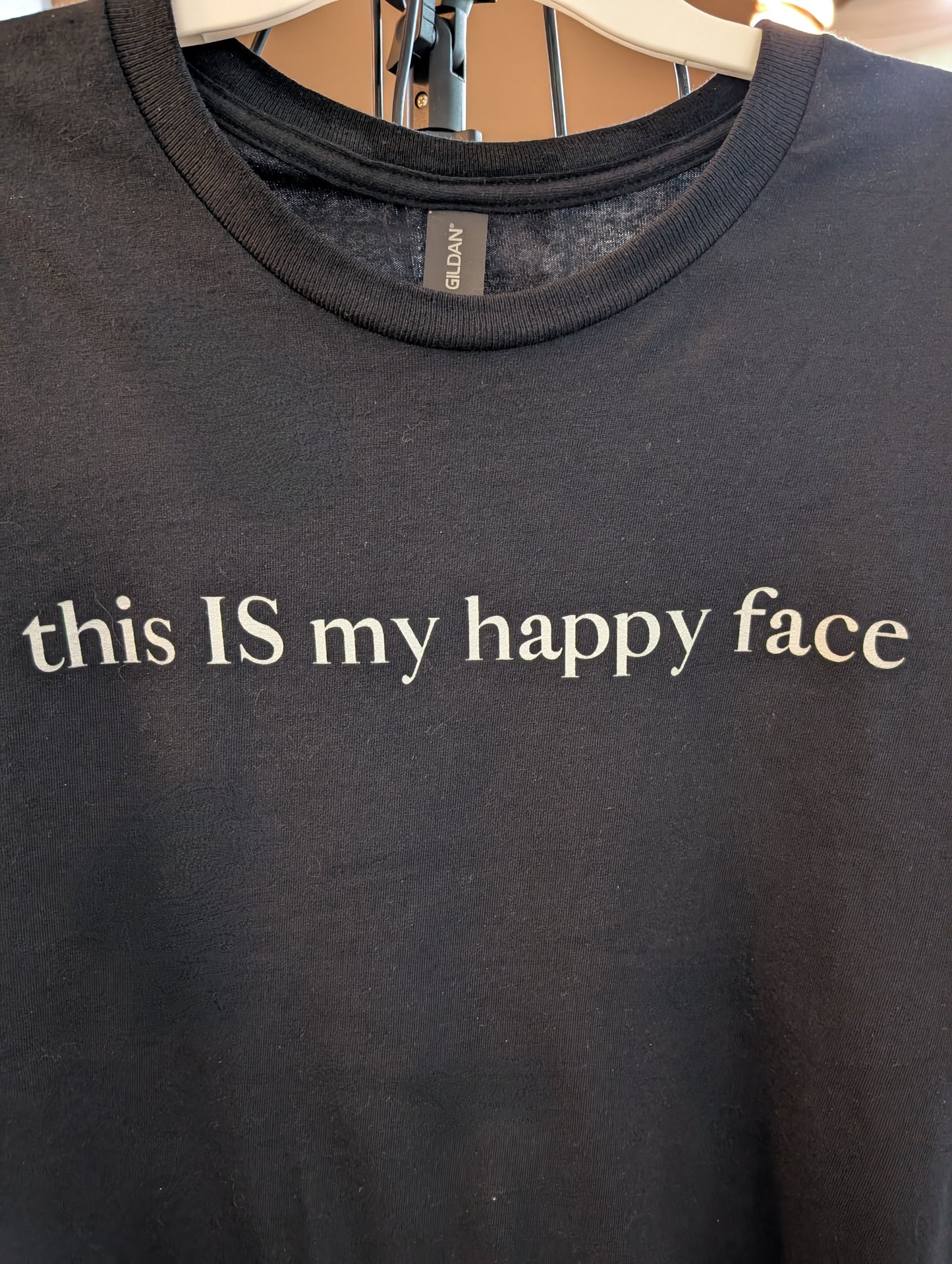 A black unisex cotton t-shirt with the text 'this IS my happy face' printed in white across the chest. All characters are lower case except the word 'IS' which are in capital letters.