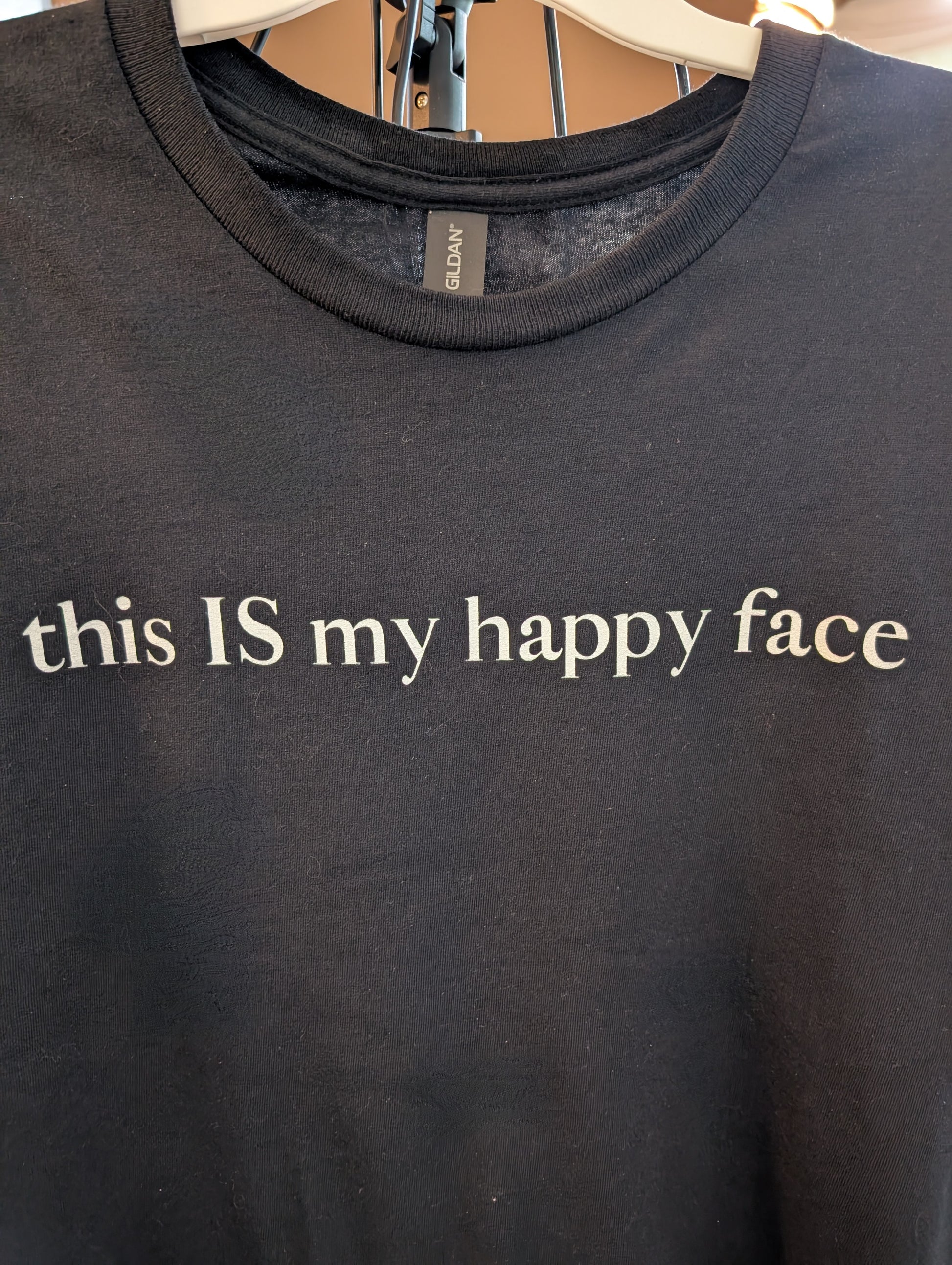 A black unisex cotton t-shirt with the text 'this IS my happy face' printed in white across the chest. All characters are lower case except the word 'IS' which are in capital letters.