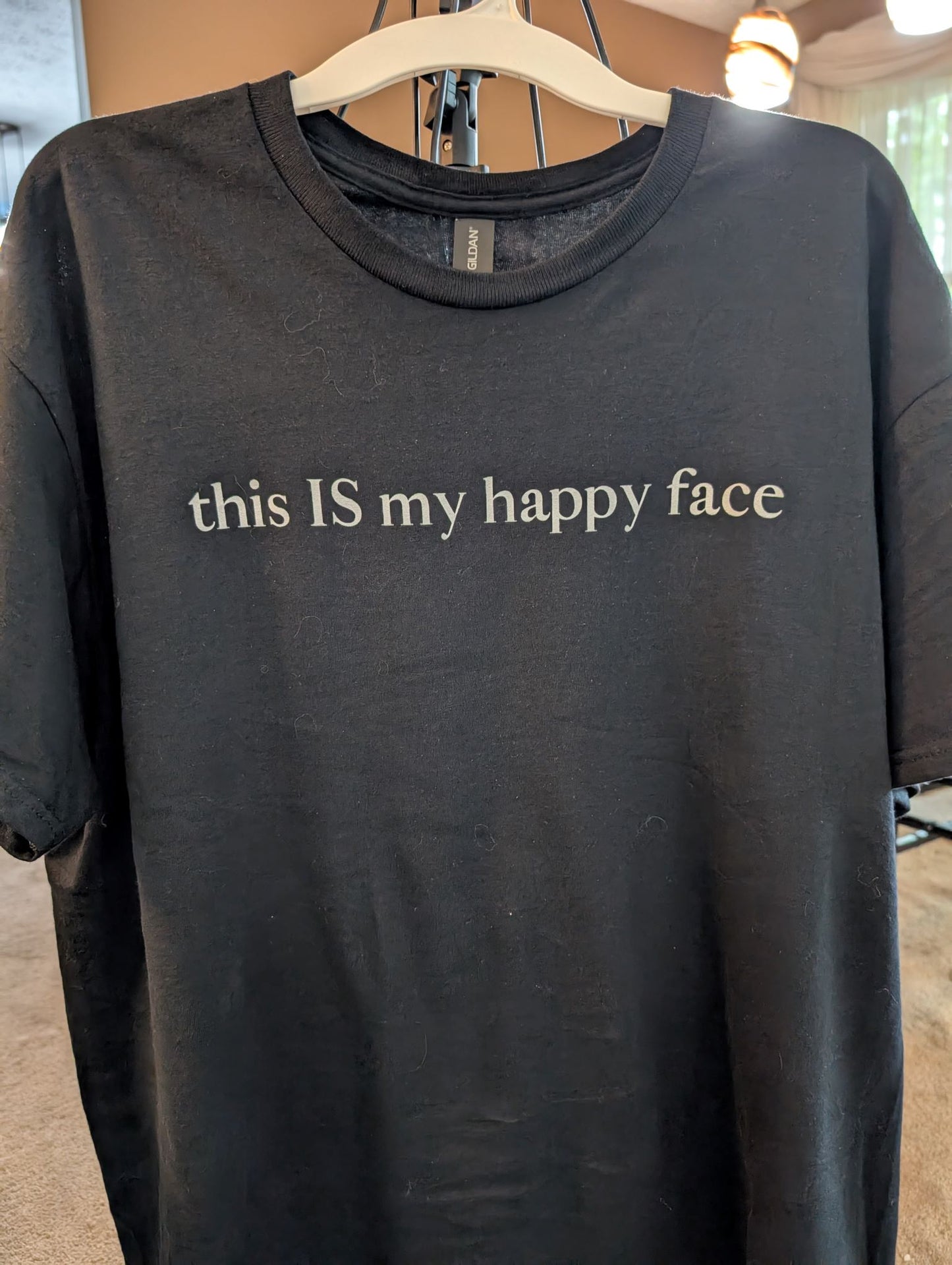 A black unisex cotton t-shirt with the text 'this IS my happy face' printed in white across the chest. All characters are lower case except the word 'IS' which are in capital letters.